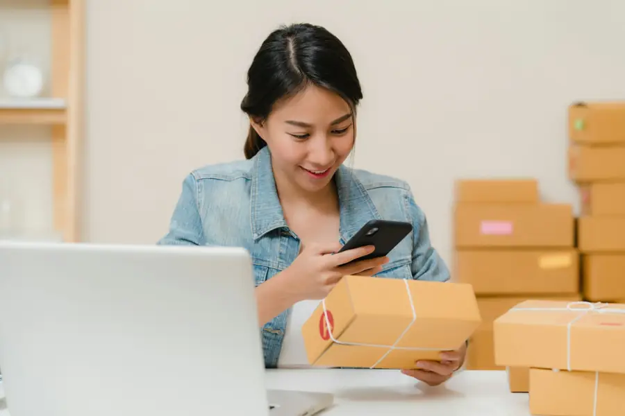 Una mujer revisa un paquete con su teléfono junto a su laptop y cajas de envío